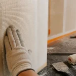 Pose toile de verre murale : main gantée maroufle la toile sur mur encollé, sans bulles